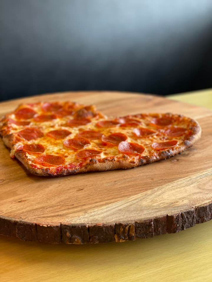 A heart shaped pepperoni pizza is sitting on a wooden cutting board.