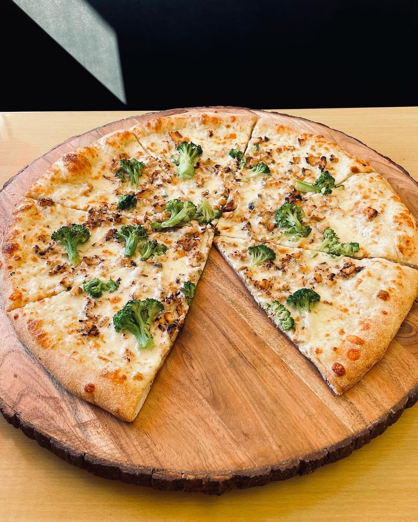A pizza with broccoli and cheese is on a wooden cutting board.