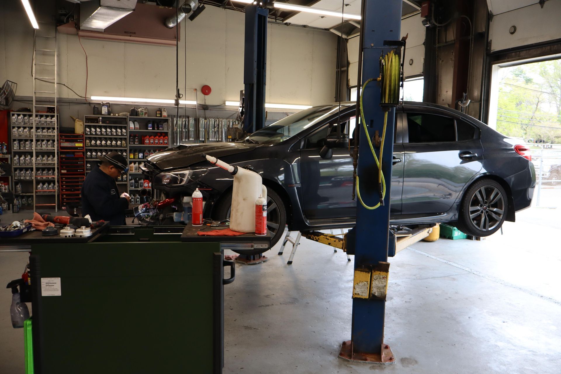 Car in auto repair shop on a lift, with mechanic working at a cluttered workbench.