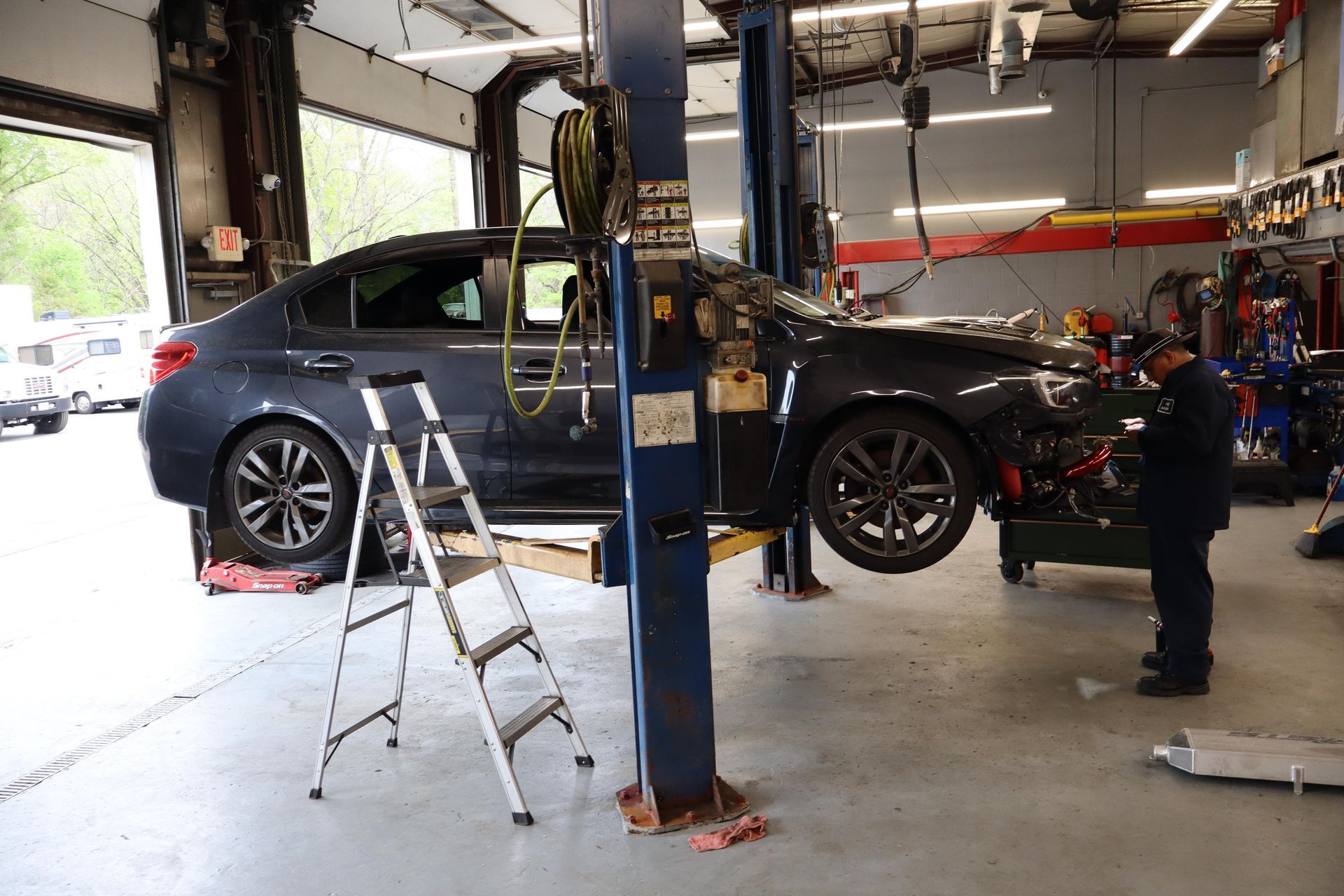 Blue car lifted in an auto repair shop, with a mechanic working nearby and a ladder in the foreground