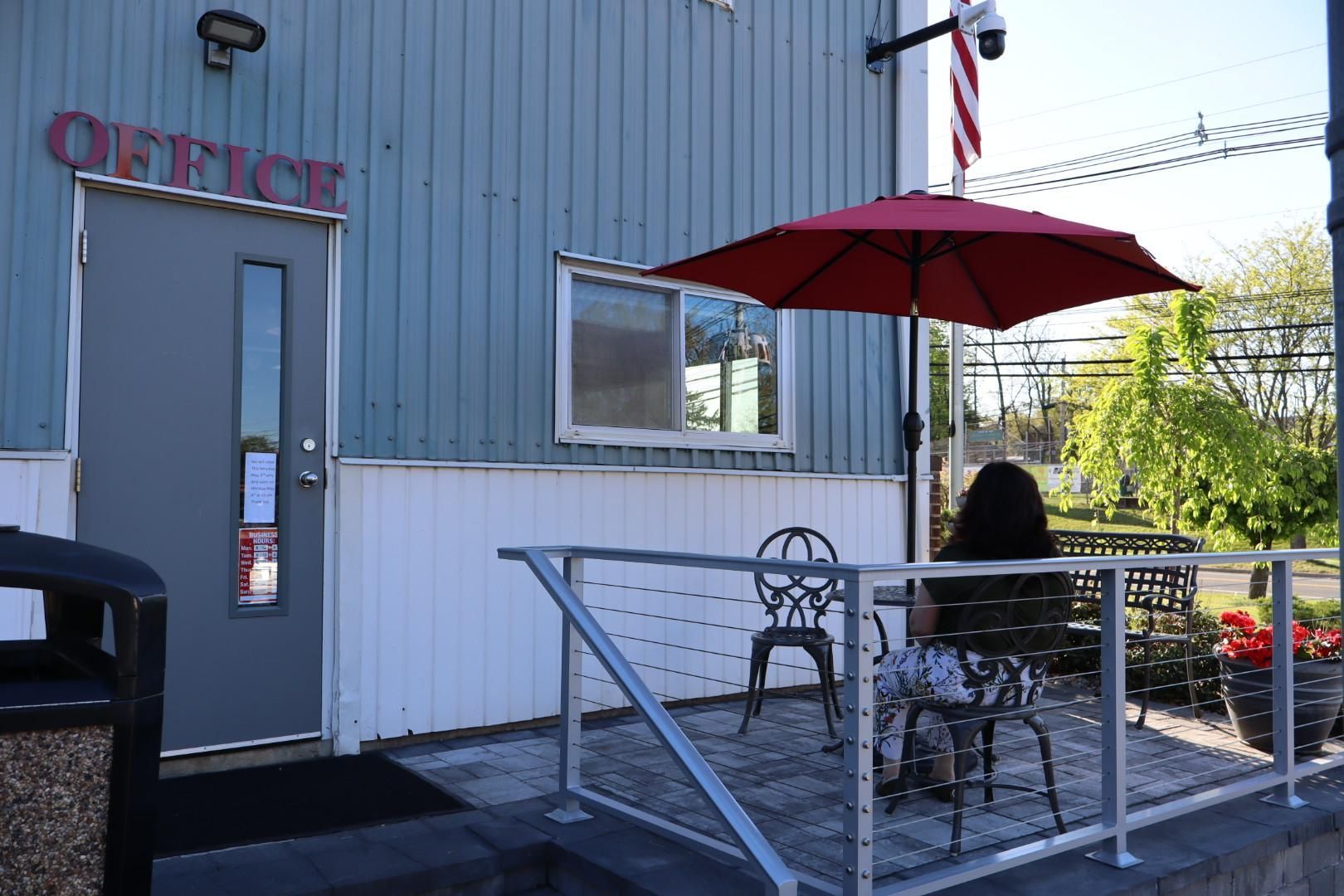 Outdoor café patio with a red umbrella, metal chairs, and a person seated by a gray building with an office door
