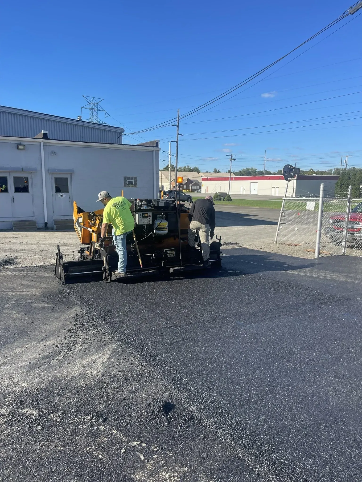 Workers paving asphalt with a machine near a building, under a blue sky.