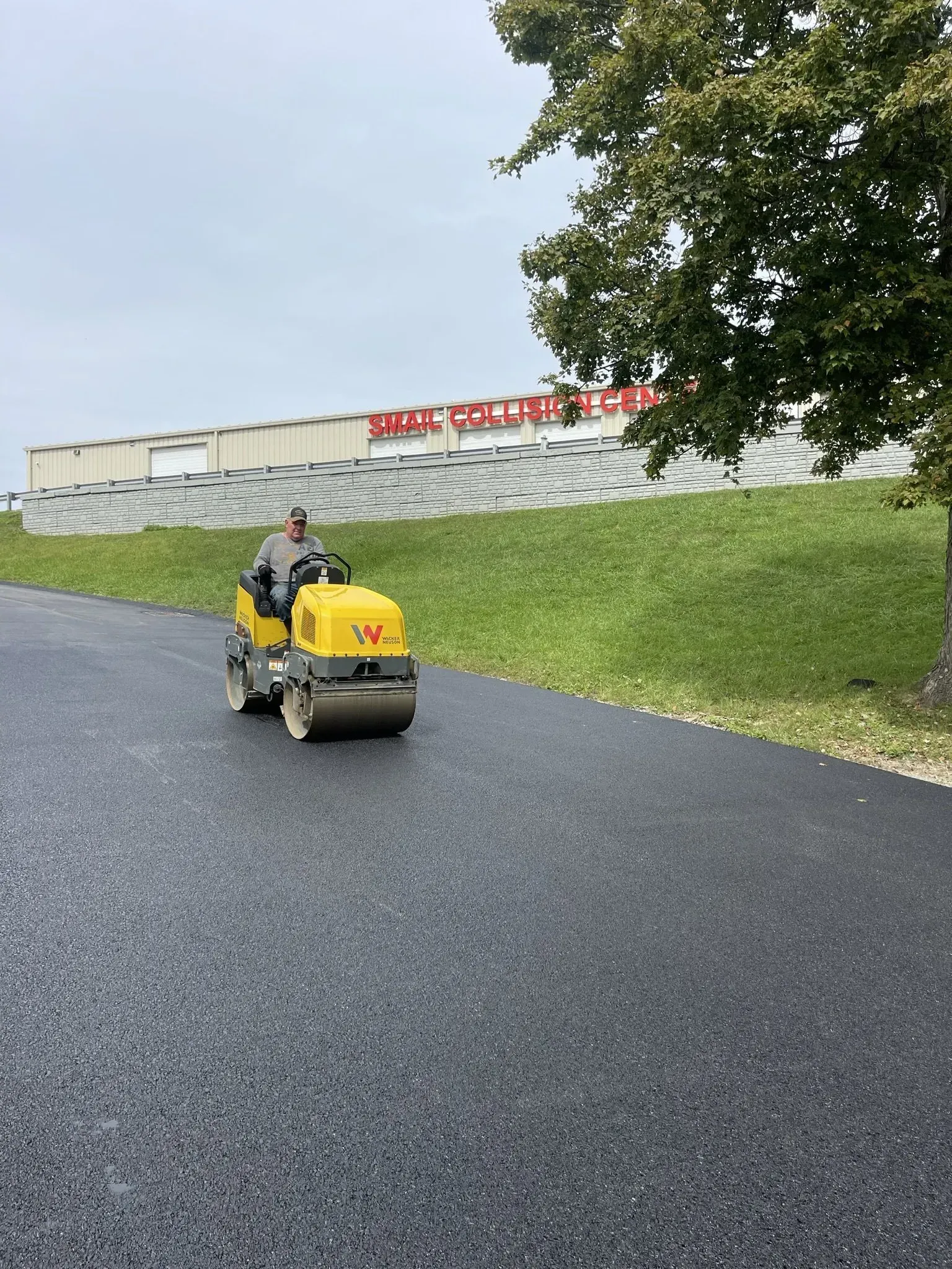 Person operating a yellow asphalt roller on newly paved road; green hillside and building in background.