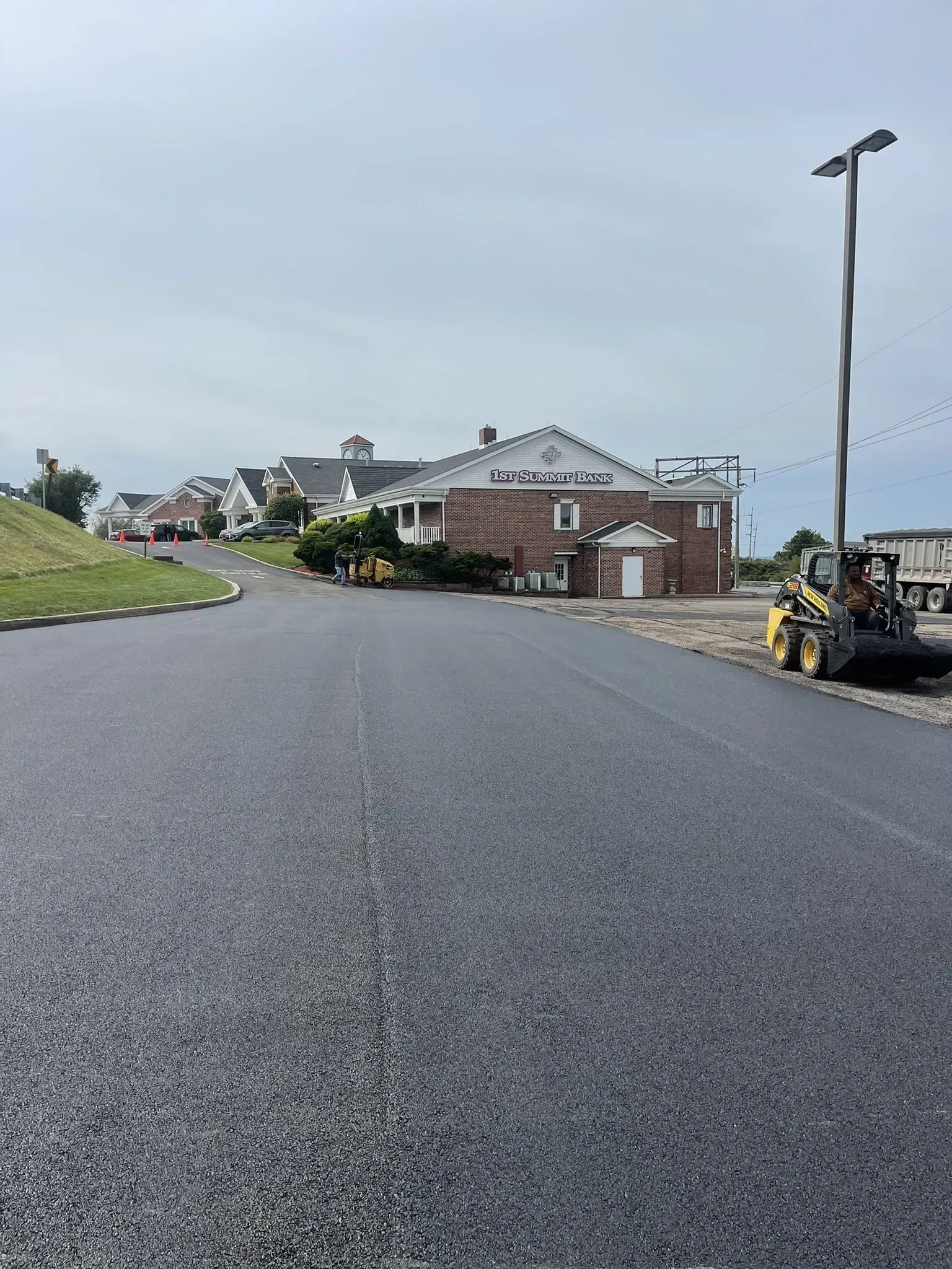 Freshly paved asphalt road leads towards buildings, a cloudy sky overhead.