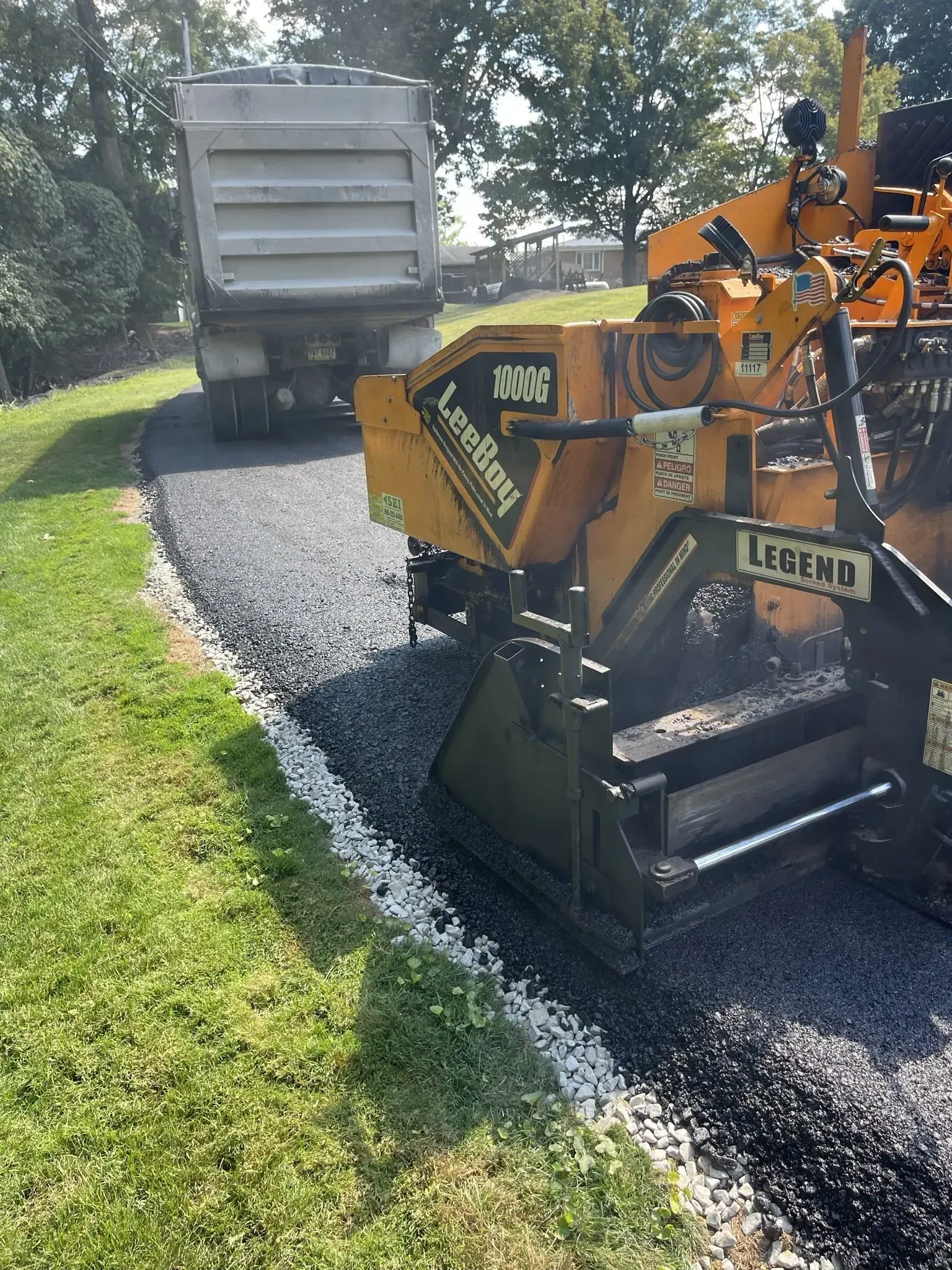 Asphalt paving machine laying black asphalt next to green grass; dump truck in background.