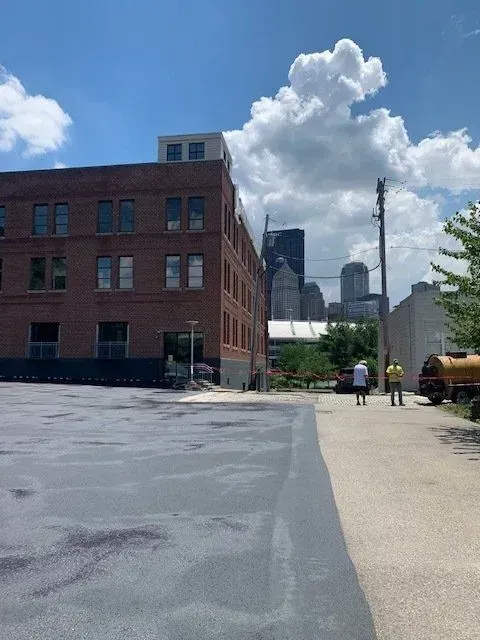Brick building next to paved area, with city skyline in the background, under a cloudy blue sky.