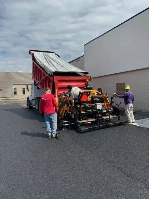 Asphalt paving in progress: truck dumping asphalt into a paver, three workers, sunny day.