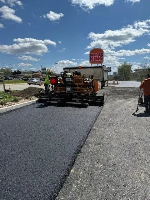 Asphalt paving in progress at a Burger King. Workers operate machinery, laying new black surface. Cloudy sky.