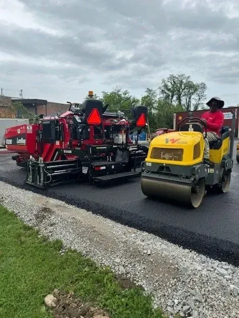 Asphalt paving in progress: Paver and roller compacting black asphalt on a road, under an overcast sky.