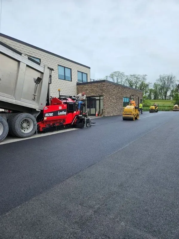 Asphalt paving in progress: truck dumping material to a paver, rollers compacting. Building in background.