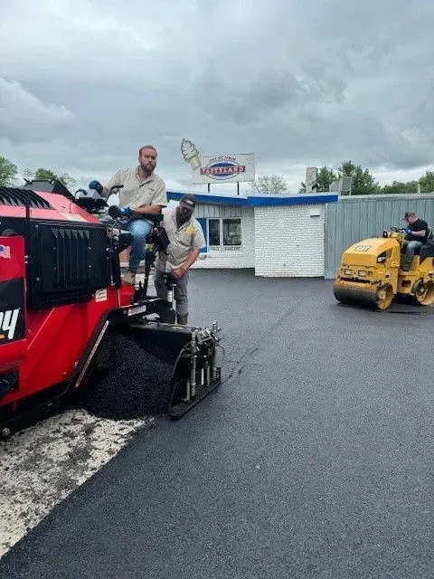 Paving crew working on asphalt. One operator on a paver, another assisting, and a roller in the background. Cloudy day.