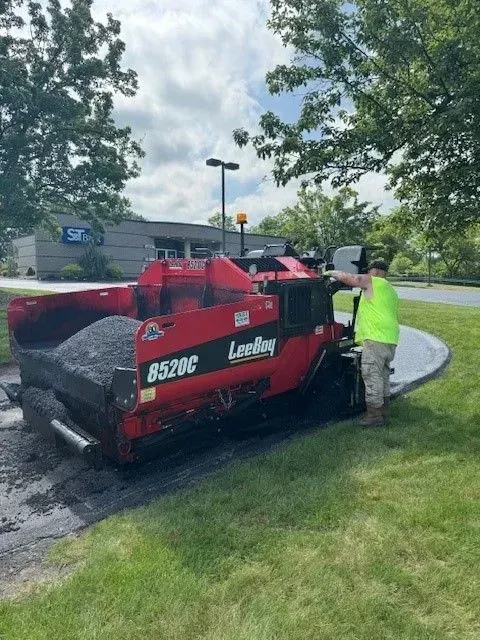 A red asphalt paving machine lays asphalt along a curved walkway. A worker in a green vest stands nearby.