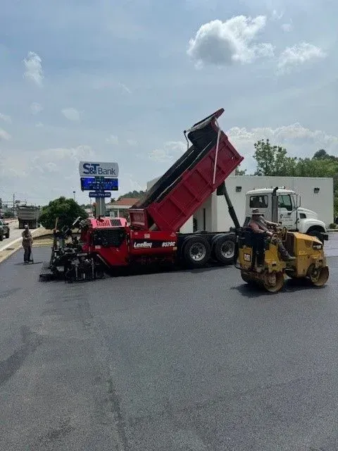 Asphalt paving: Dump truck unloading onto a paver. Roller compacting fresh asphalt; a worker stands by.