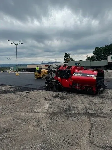 Red asphalt paver laying asphalt; worker operating small yellow roller. Cloudy sky, commercial setting.