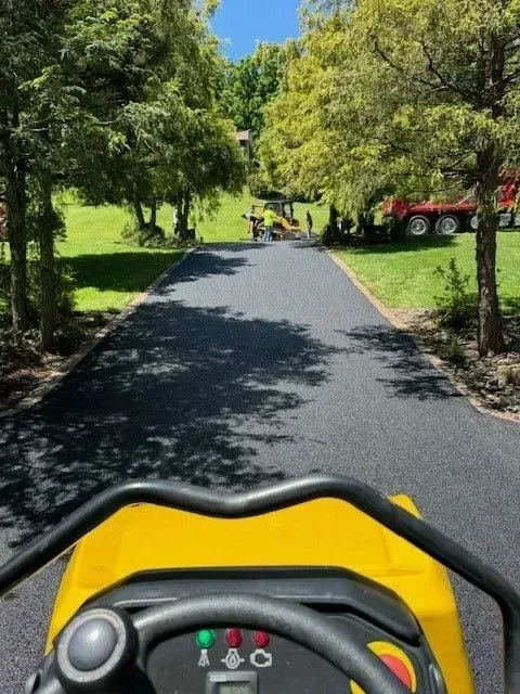 A person operating a yellow asphalt compactor on a newly paved road bordered by trees and grass, with construction equipment visible.