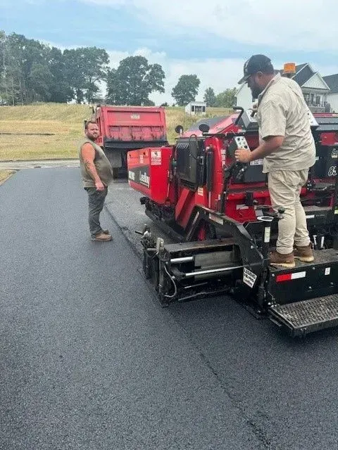 Workers paving a road with asphalt. One operates the paver, the other observes by truck.