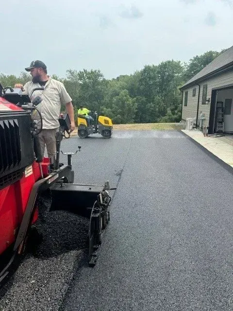 Asphalt paving in progress: man operates a red machine, another on an ATV sprays material.