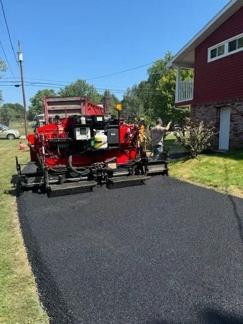 Asphalt paving machine laying black asphalt on driveway; person guiding. Red building in background.