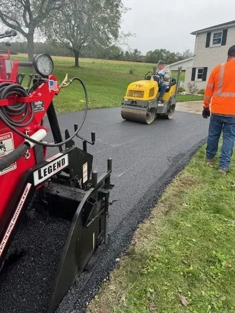 Asphalt paving of a driveway, with a Legend paver, roller, and worker in orange vest.