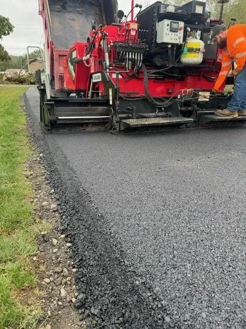 Road paving machine laying asphalt on a road, worker in orange vest.