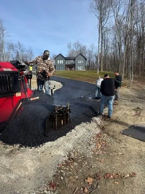 Workers paving a driveway with asphalt near a two-story gray house.