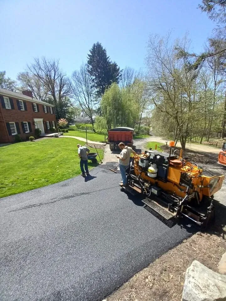 Asphalt paving in progress: workers operating a paving machine on a driveway. Truck in the background. Sunny day.
