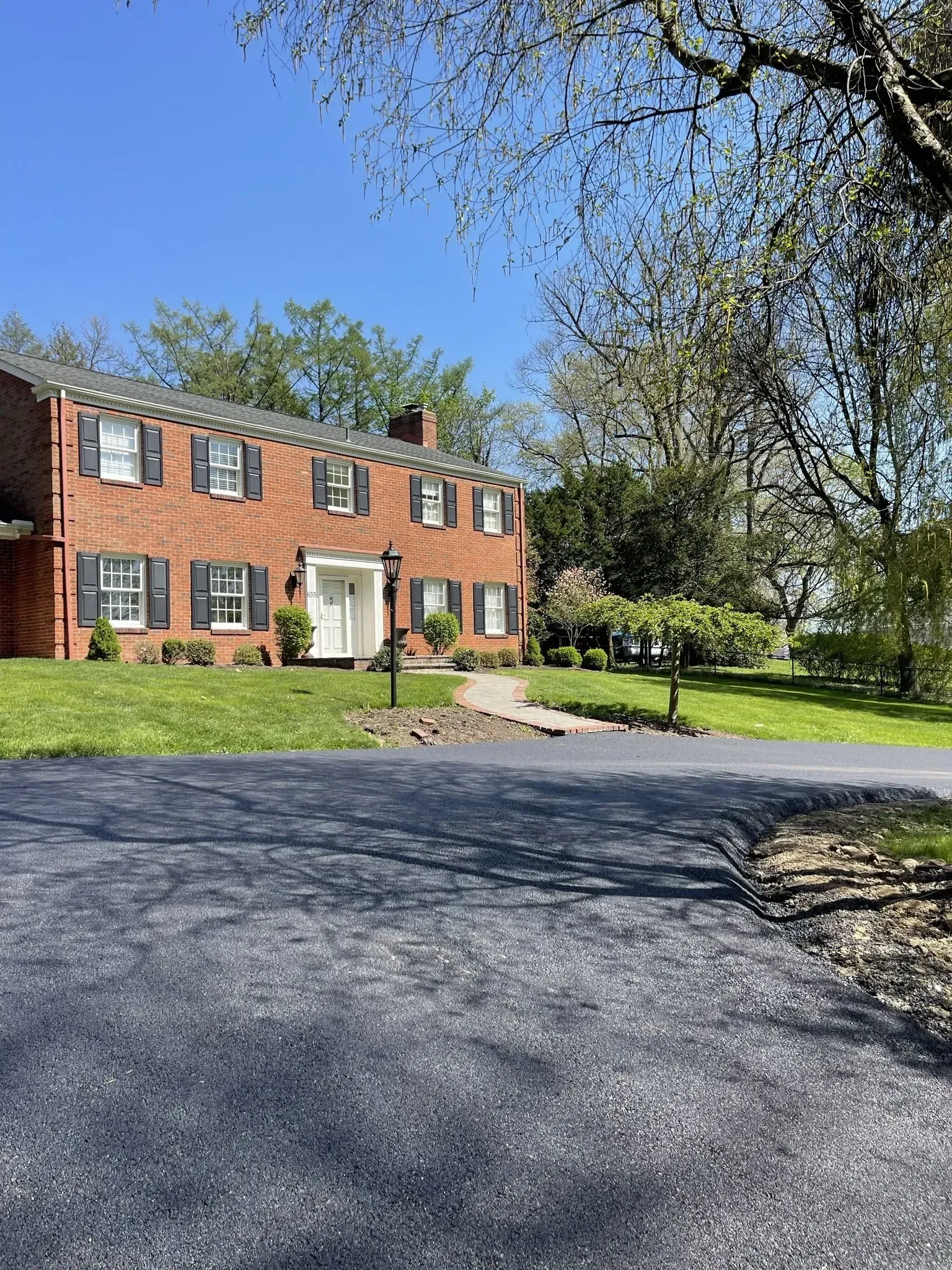 Brick two-story house with black shutters, white door, and a black paved driveway. Trees and green grass surround.