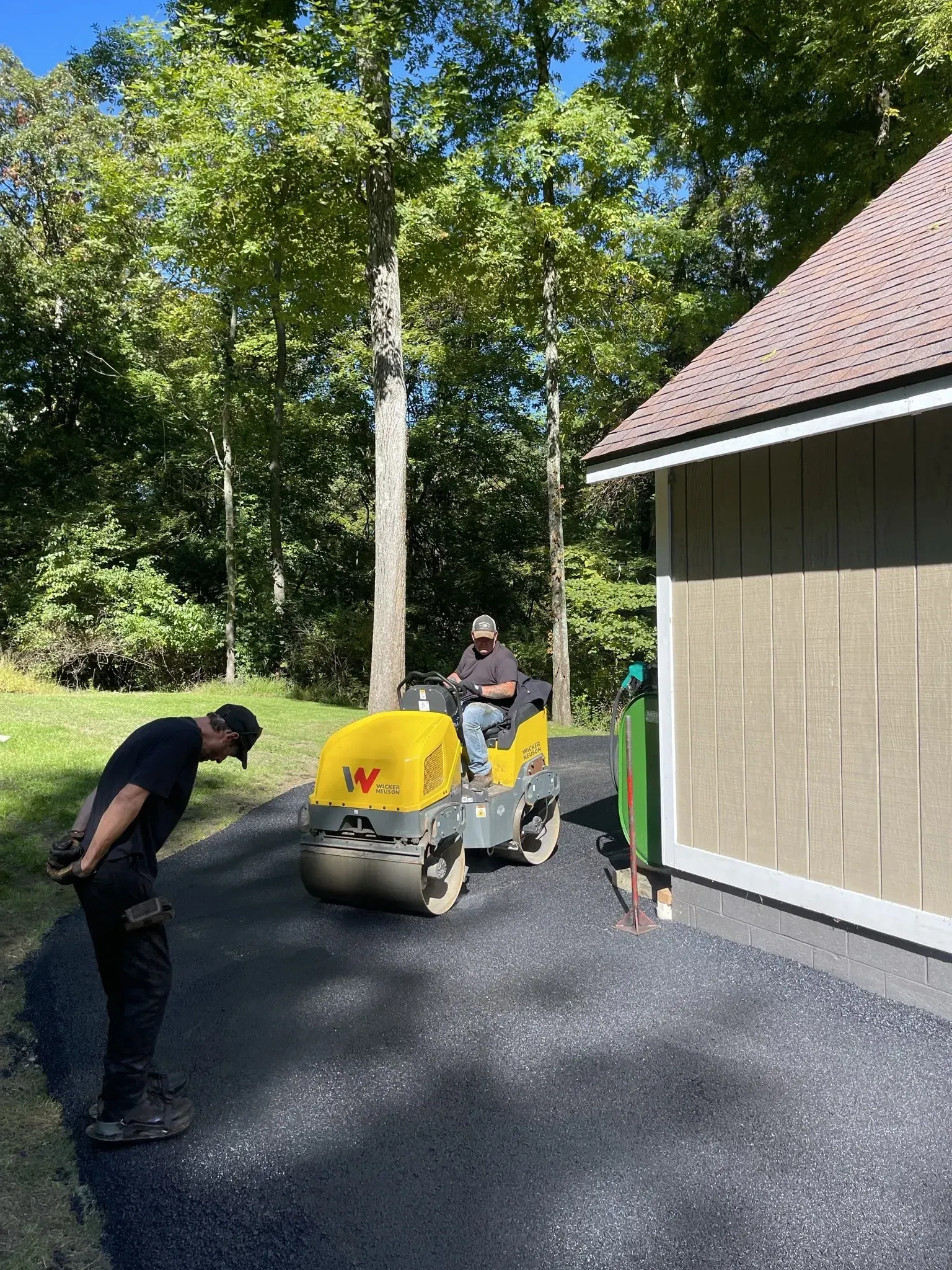 Man operating a yellow roller compacting asphalt on a driveway as another man watches.