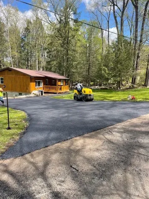 Asphalt driveway being rolled by a small machine in front of a wooden house with a metal roof.
