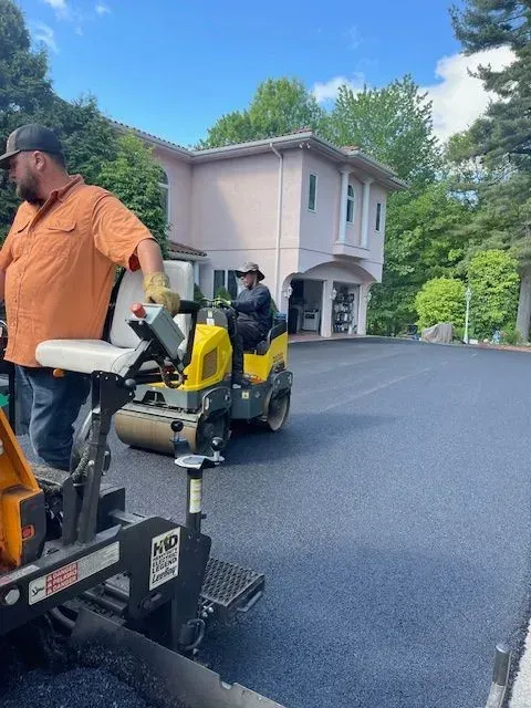 Road crew paving a driveway with asphalt roller in front of a light-colored house.