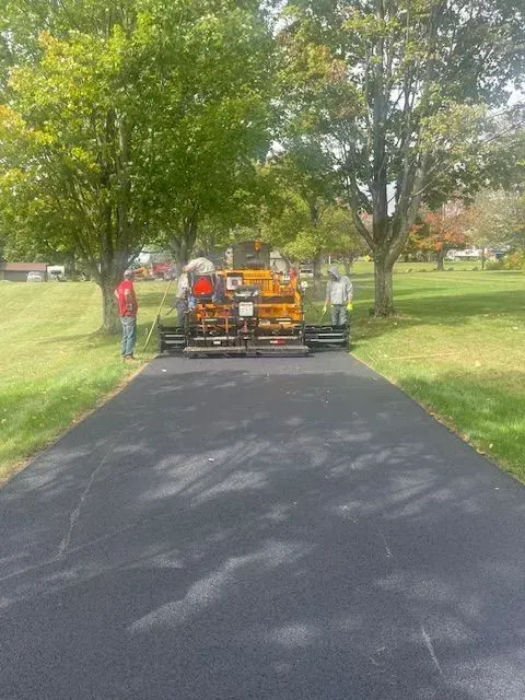 Paving crew operating machinery on a newly paved asphalt road, with trees and grass in the background.