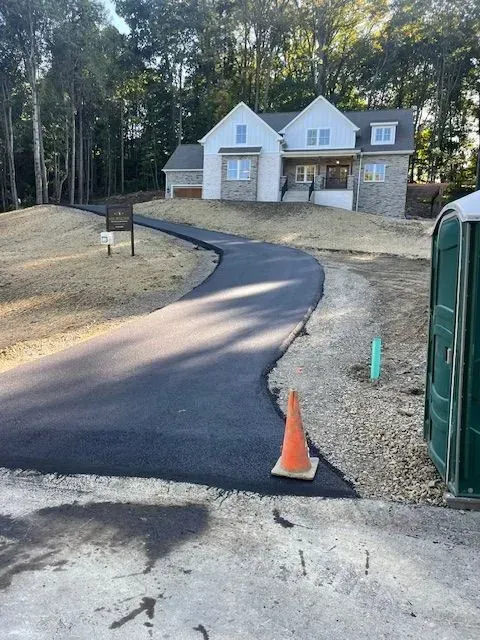 Newly paved asphalt driveway leading to a two-story house with stone accents, orange traffic cone present.