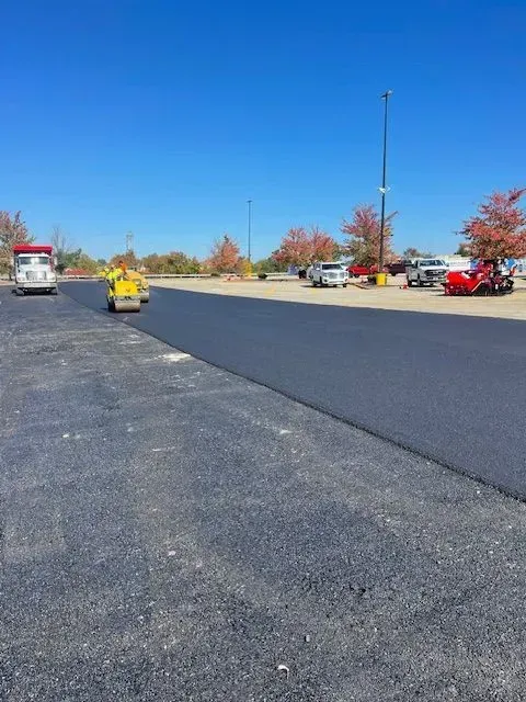 Parking lot paving in progress; asphalt roller, trucks, cars, trees, bright blue sky.