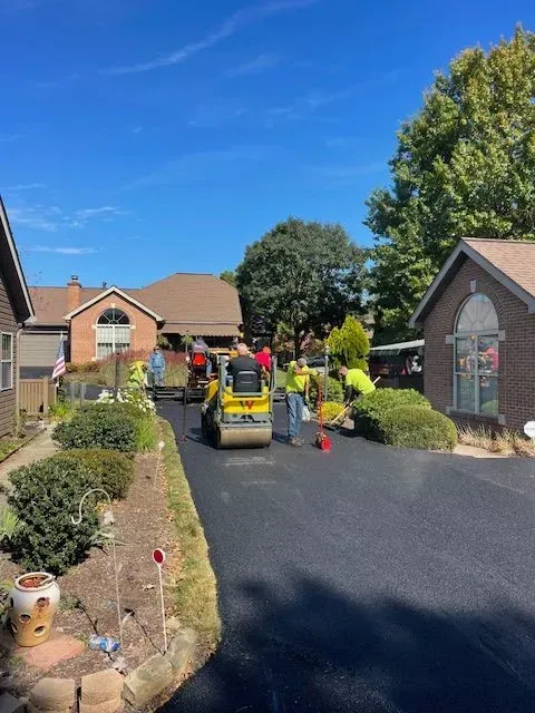 Asphalt paving crew working on a driveway in front of suburban houses. A roller compacts the fresh black asphalt.