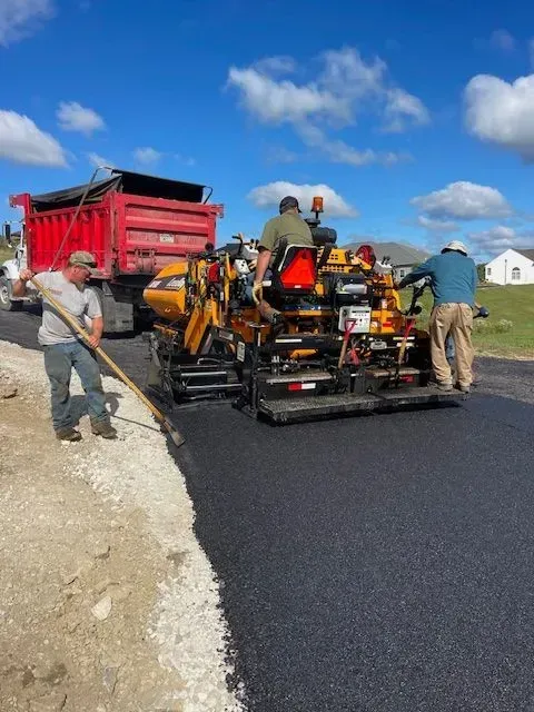 Asphalt paving in progress: workers operating a machine, truck delivering asphalt, bright sky.