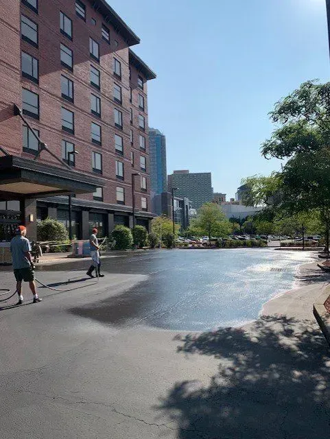 Two people washing a wet asphalt area near a brick building. Trees and other buildings are in the background under a blue sky.