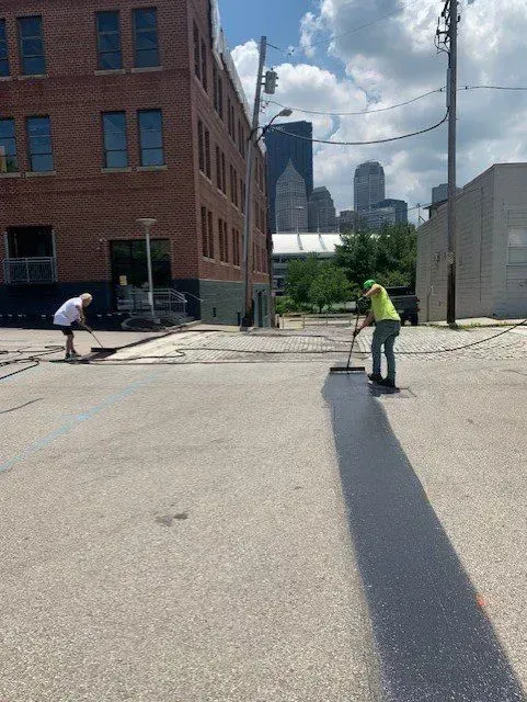 Two people seal a section of asphalt on a city street; brick building in background.