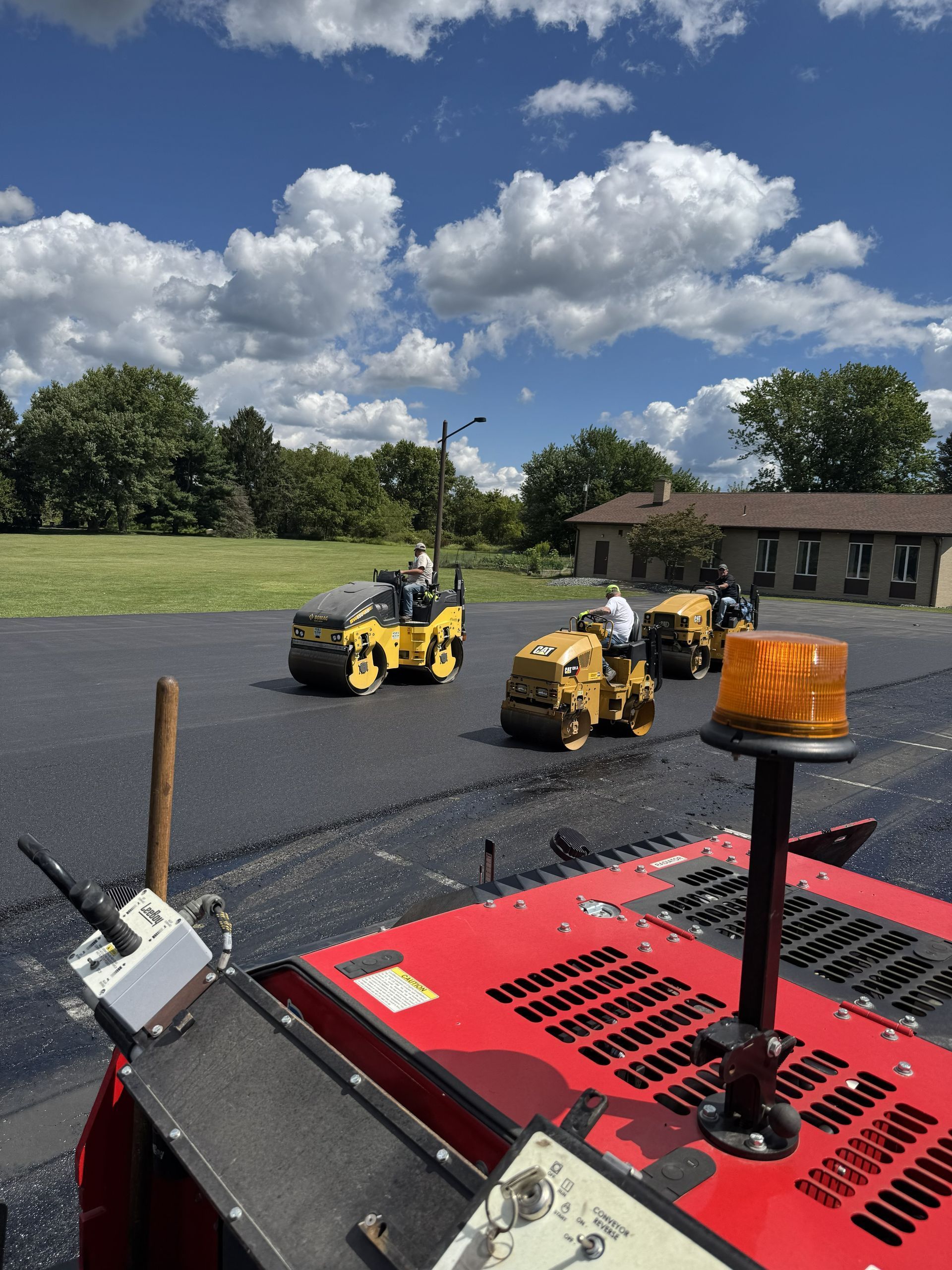 Asphalt paving in progress; three yellow rollers on fresh blacktop under a blue sky.
