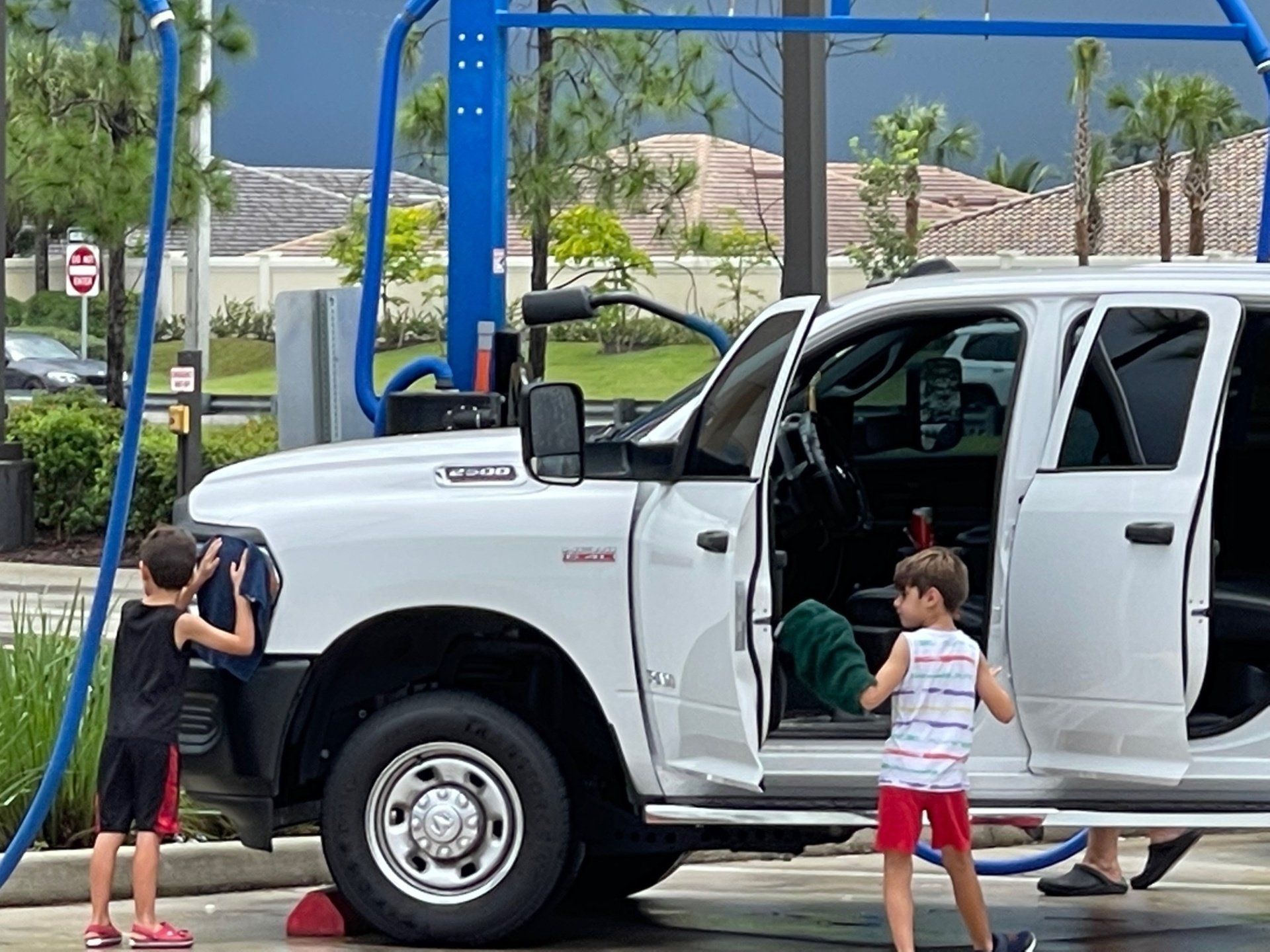 Two boys washing a white truck at a car wash; a blue hose and a cloudy sky in the background.