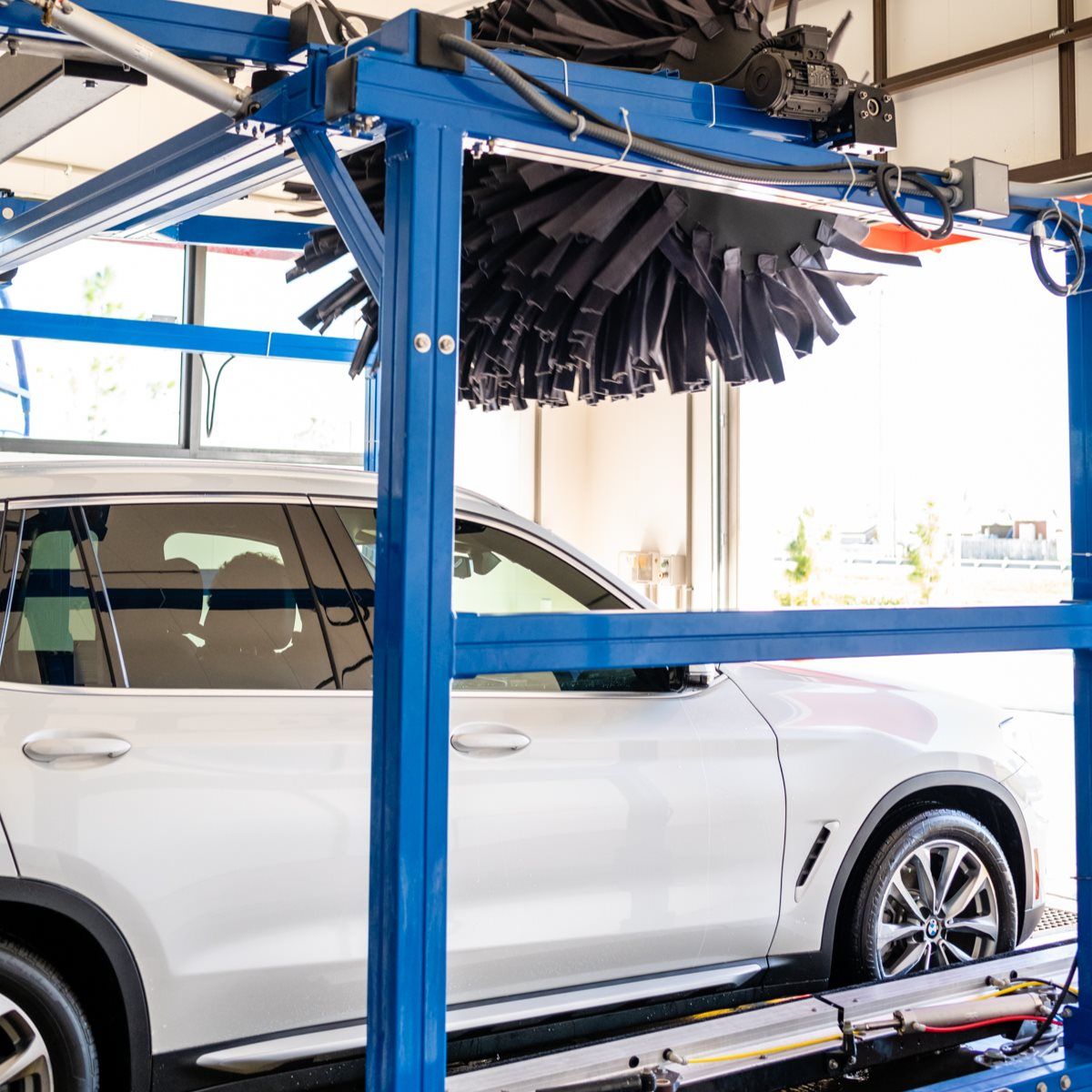 White SUV in a car wash, blue frame, black brushes overhead.