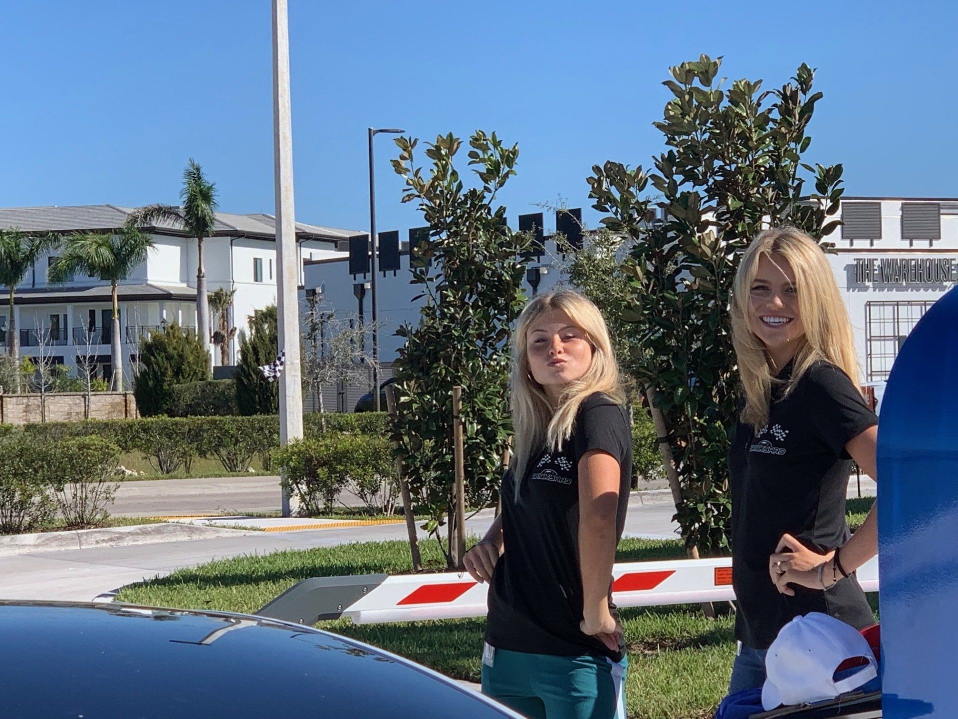 Two smiling young women in black t-shirts and sunglasses near a car. Sunny outdoor setting.