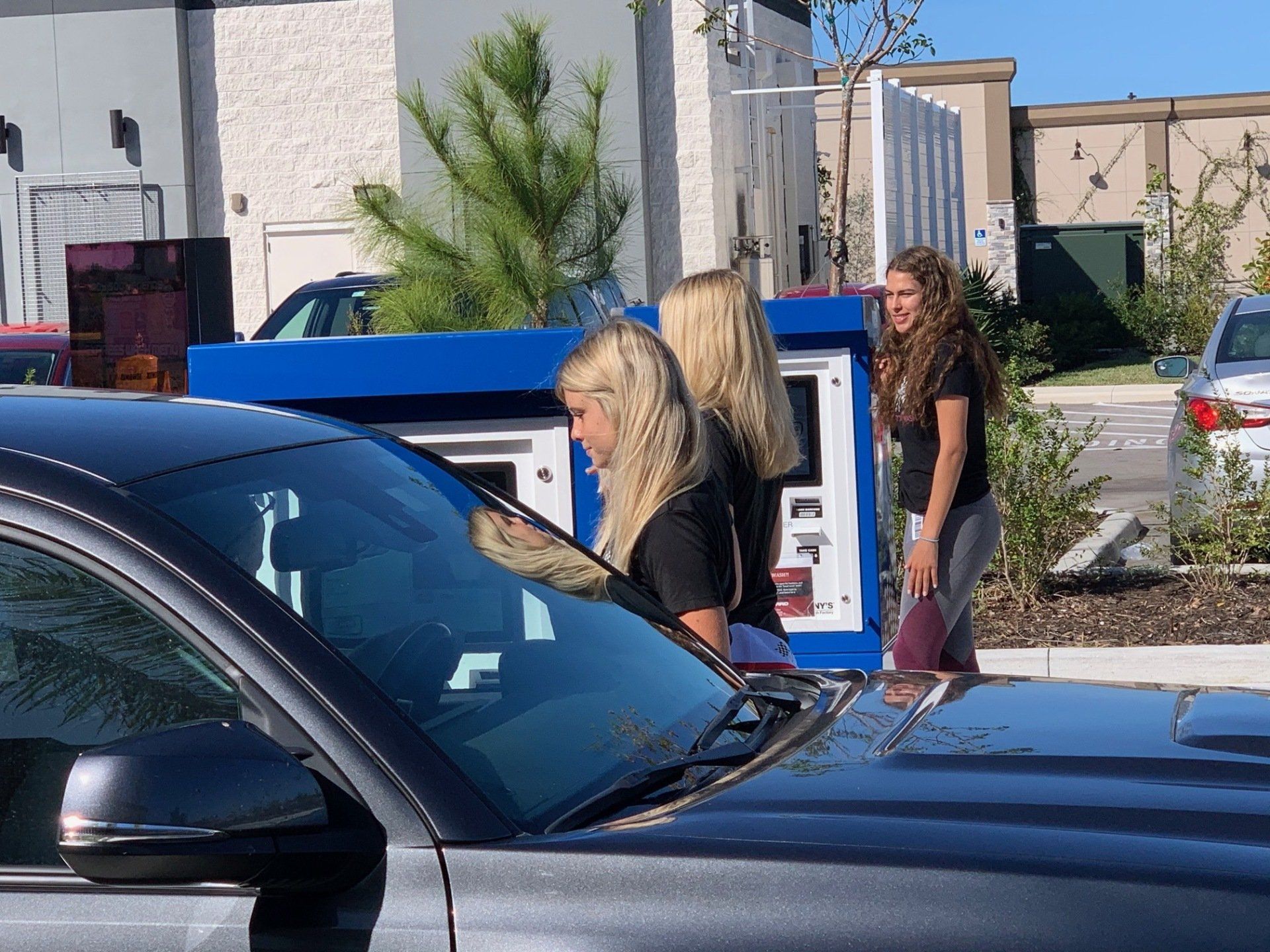 Three young women at a car wash, two blondes at the machine, one smiling.