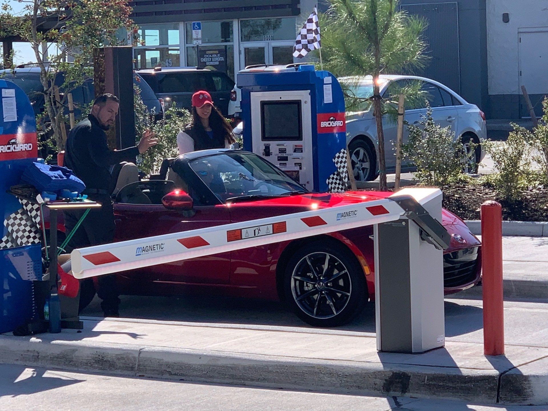 Red convertible at car wash entrance, people interacting with kiosk, barrier arm up.