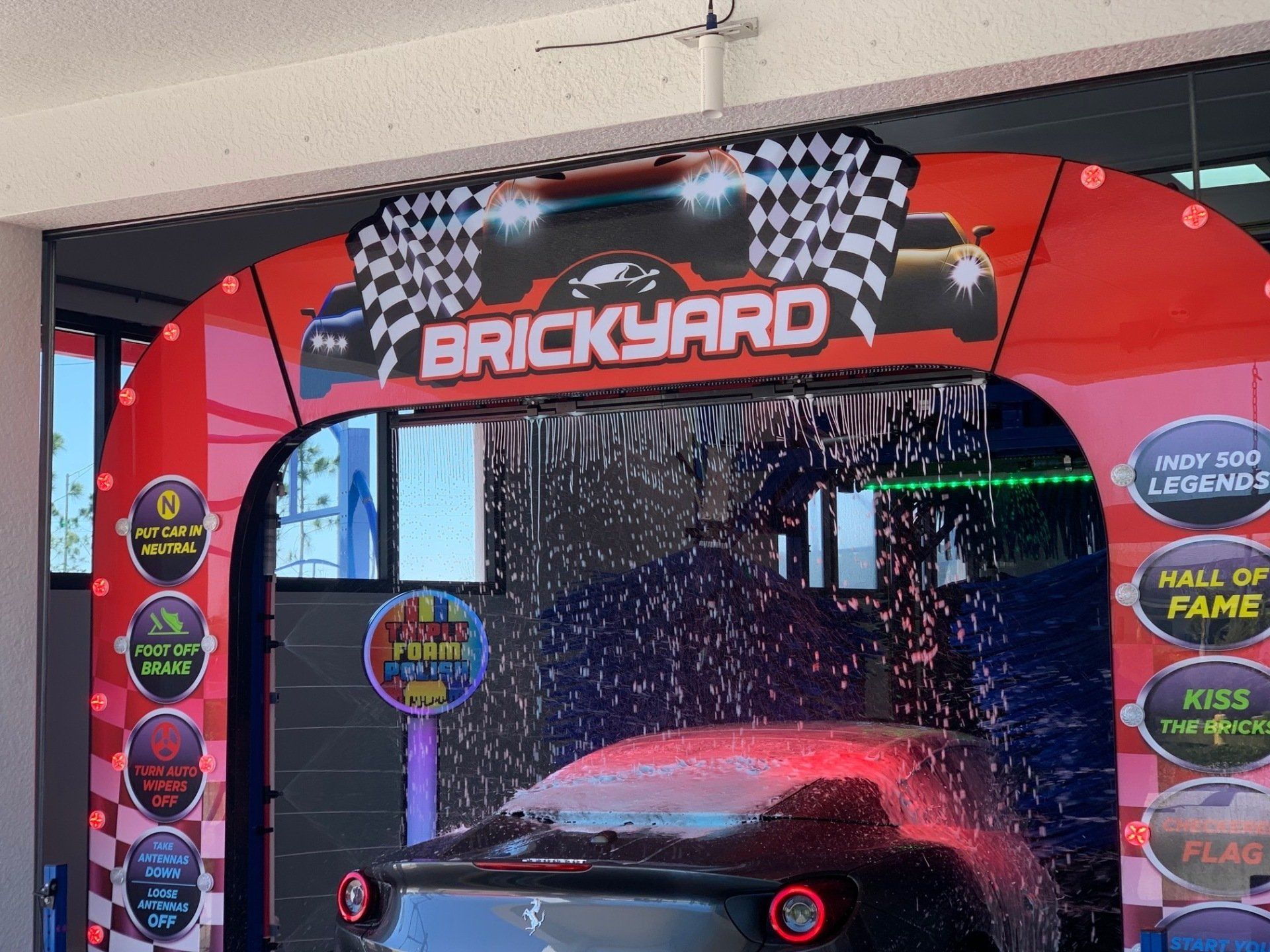 A black car inside a Brickyard car wash. Red frame, checkered flags, water spraying, and colorful lights.