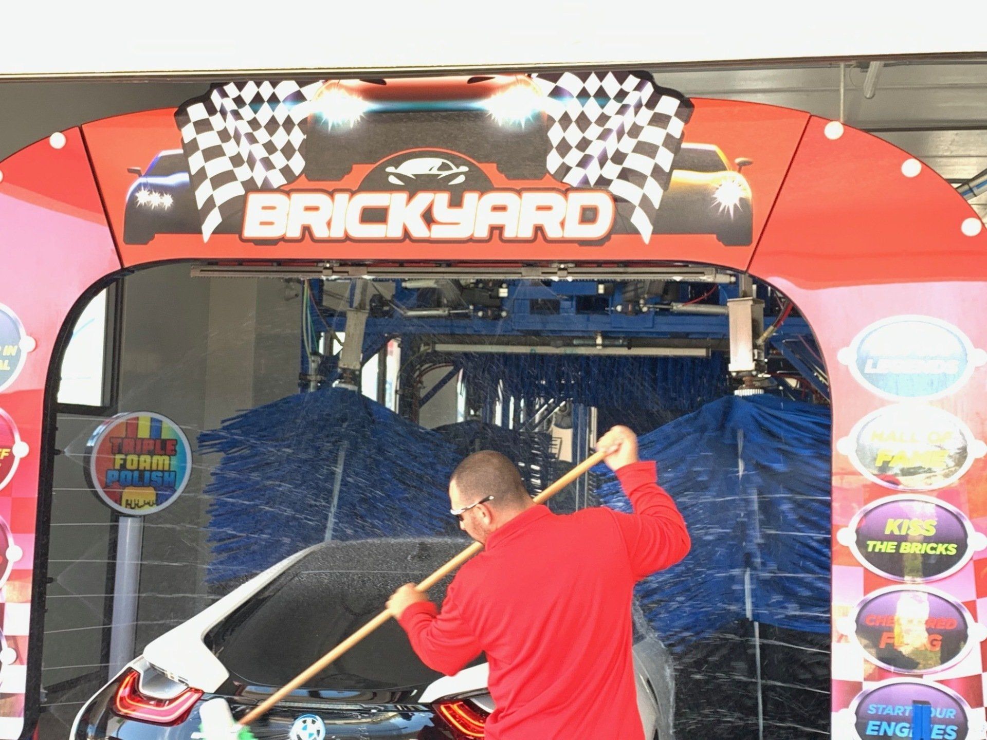 Man in red shirt pre-washes a car at the Brickyard car wash, using a long-handled brush.