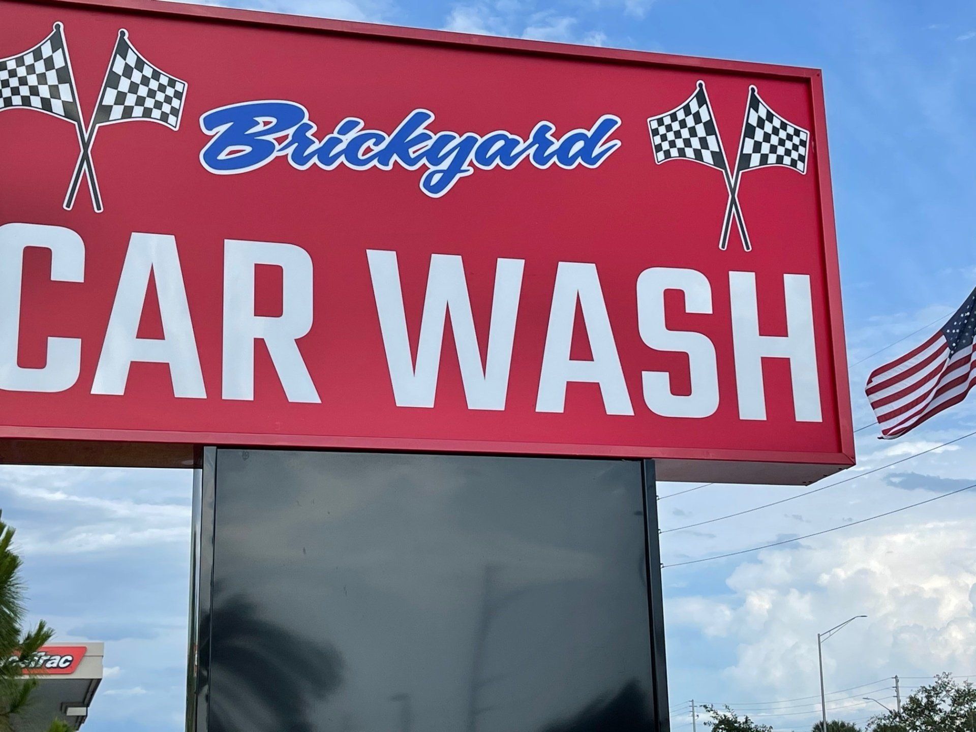 Red sign for Brickyard Car Wash with checkered flags and a black display panel; American flag in background.