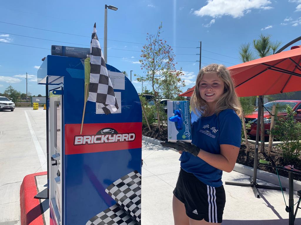 Young woman in blue shirt, black shorts, and gloves, at Brickyard carwash kiosk, holding a blue item.