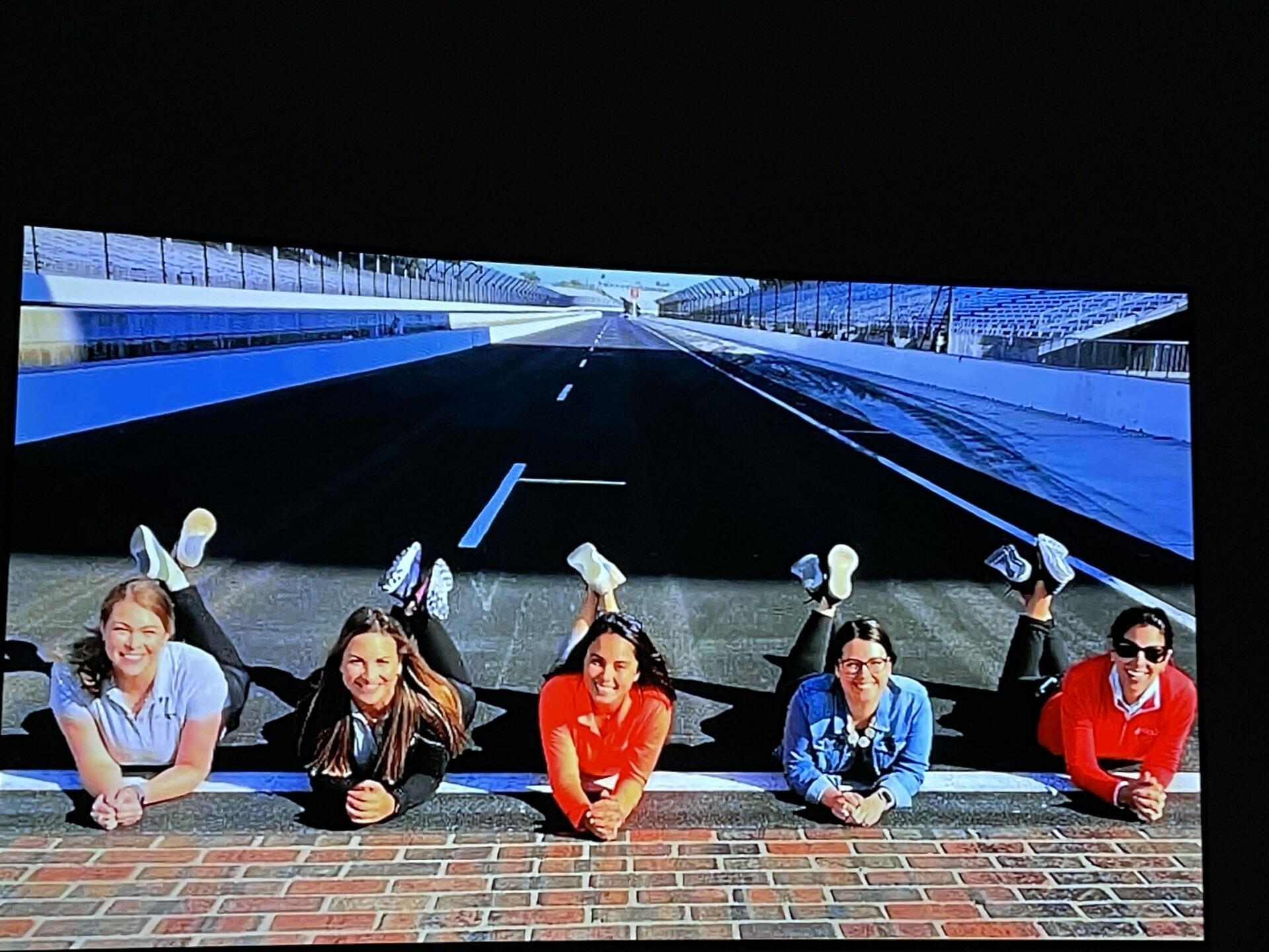 Five people lie on a brick ledge, posing with a road background. They smile and wear casual clothes.