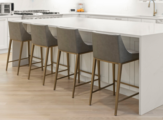 Four gray upholstered bar stools with gold legs at a white kitchen island. Light wood flooring.