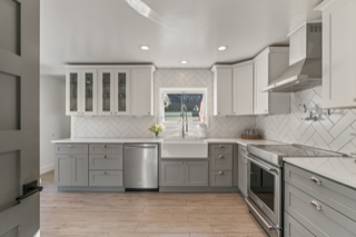 Modern kitchen with gray and white cabinets, stainless steel appliances, and a farmhouse sink. Light wood floors and white backsplash.